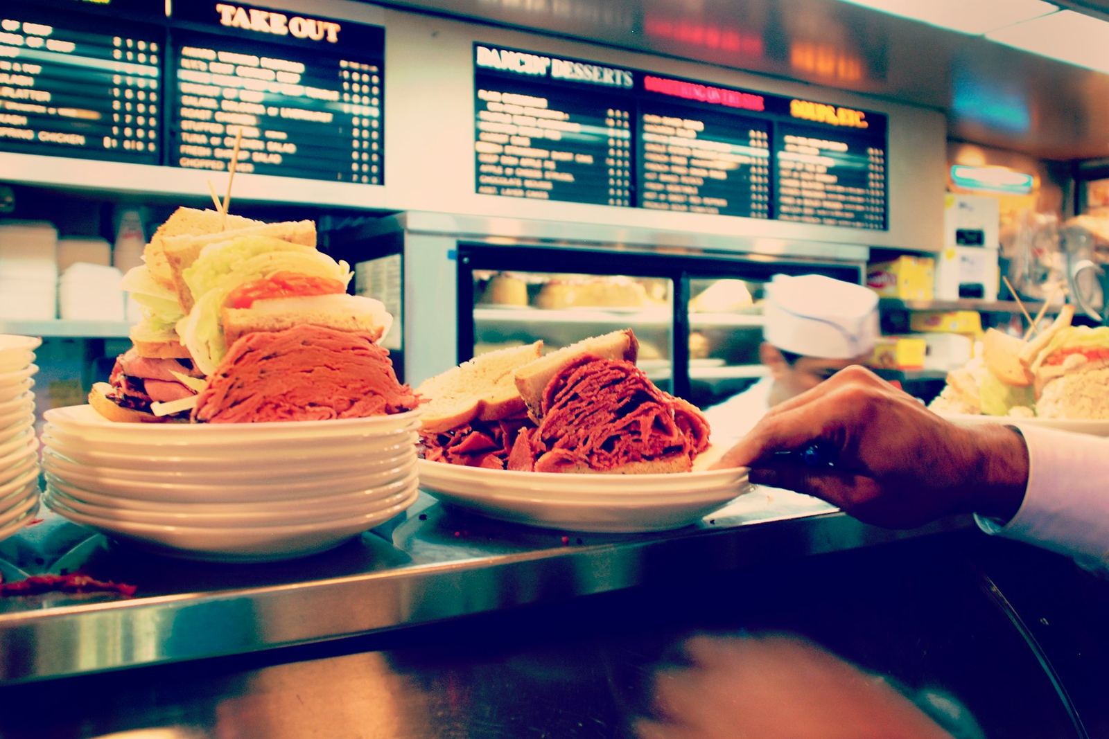 Counter service at a deli: stacked sandwiches on plates and a menu board that reads Take Out