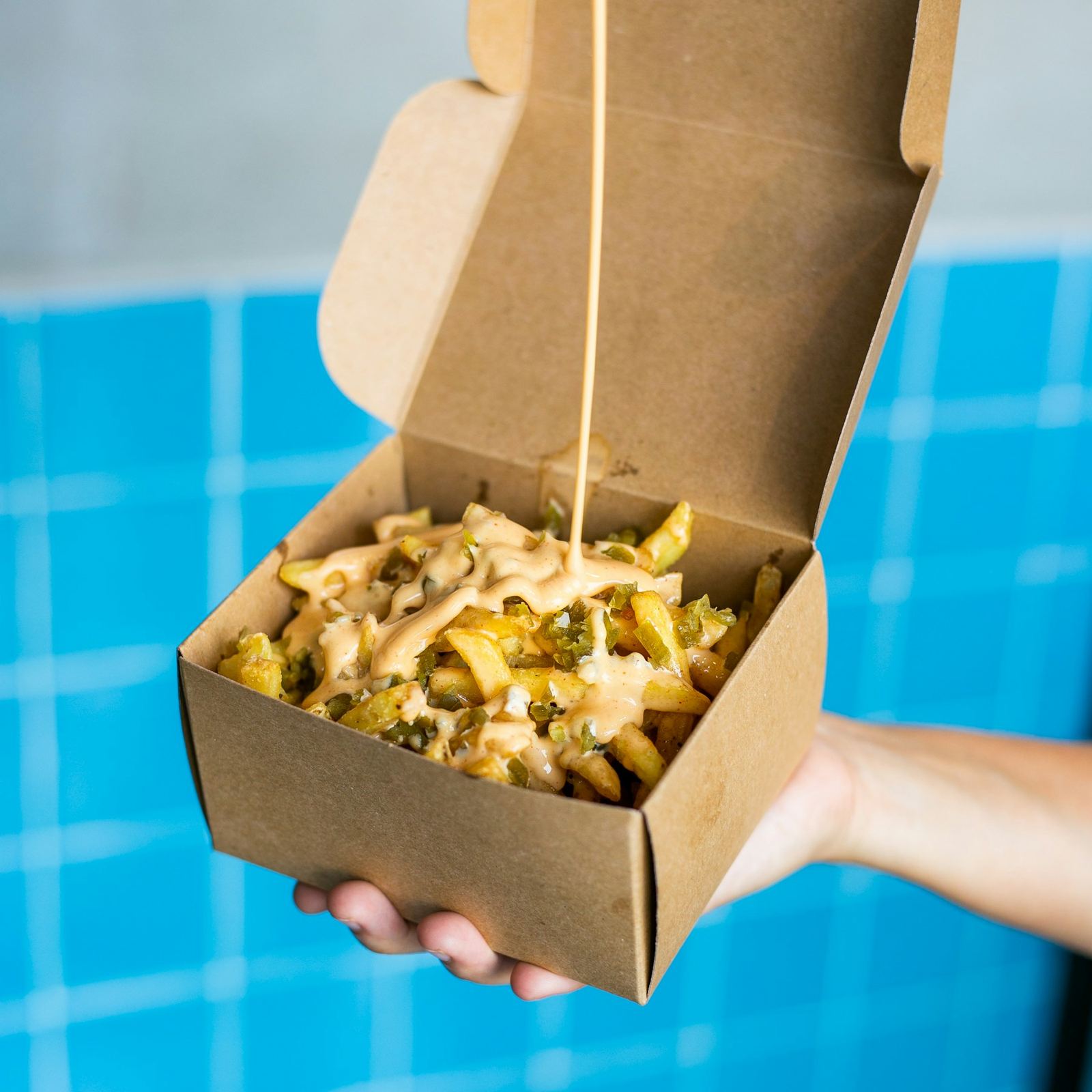 A hand holding a cardboard box of loaded fries with a drizzle of sauce