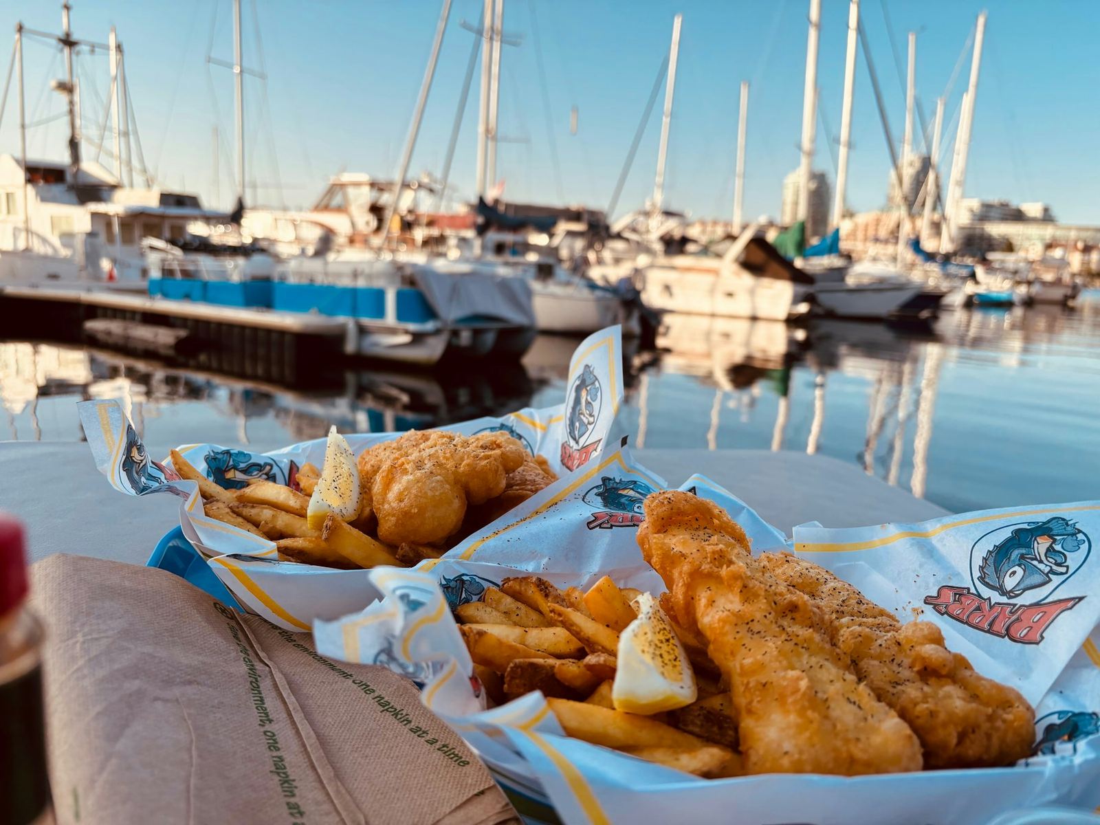 Two paper boats of fried fish and chips on a table in front of a marina full of sailboats